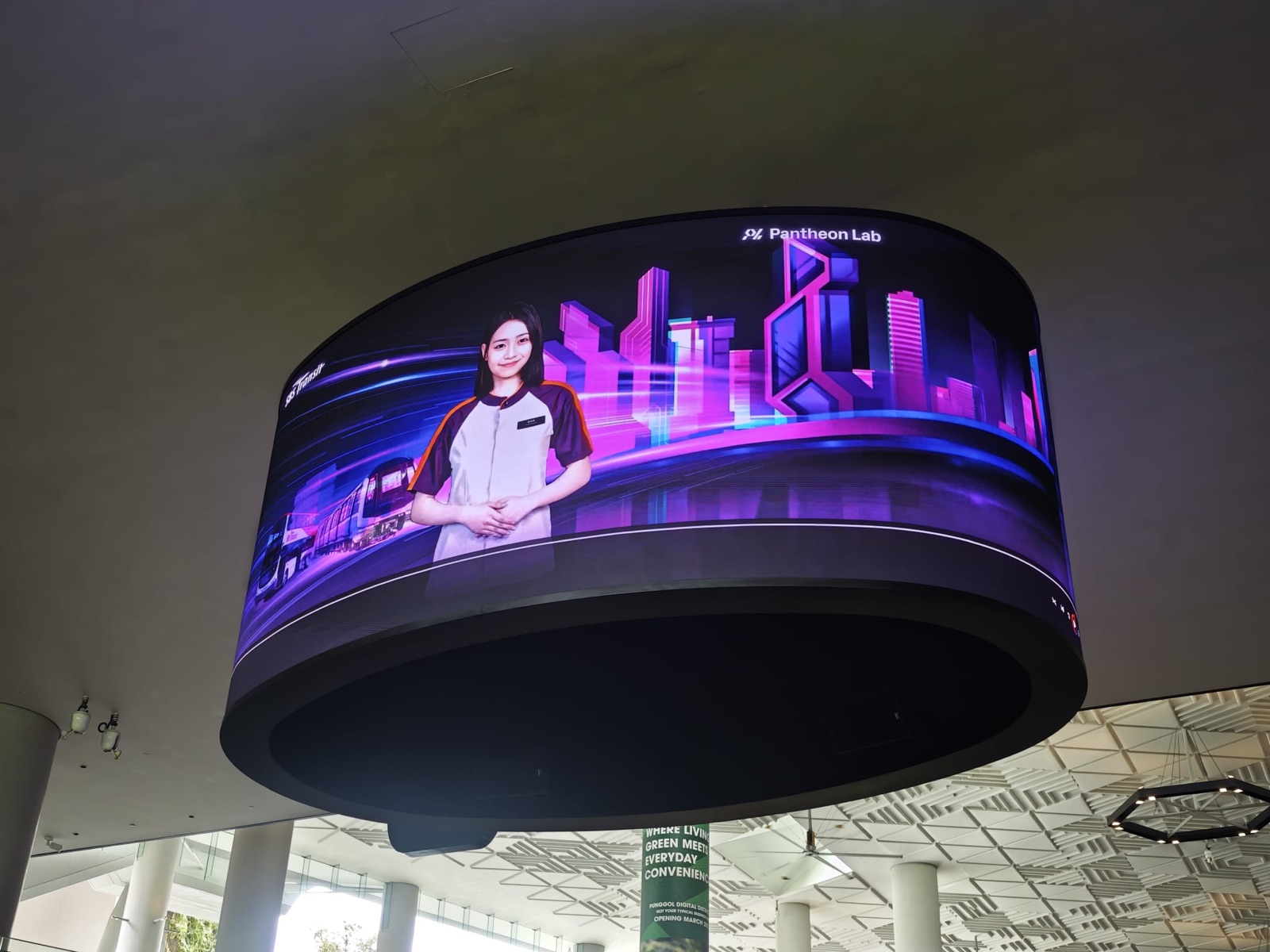 AIVA, in SBS Transit uniform, on a cylindrical LED screen suspended above a concourse, set against a neon city skyline.